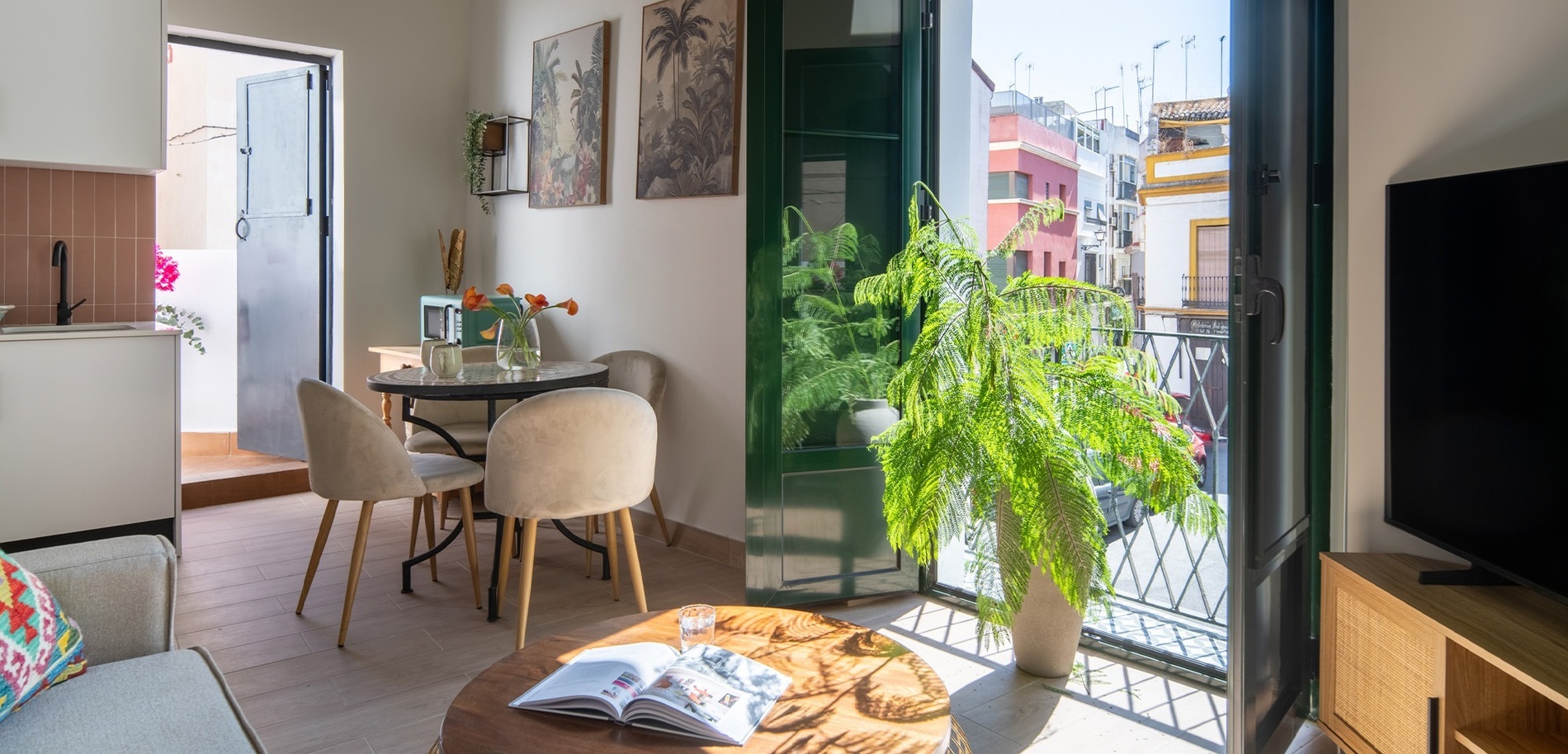 Un salón-comedor luminoso y moderno con una cocina abierta, un balcón con una planta exuberante y vistas a una calle urbana colorida.