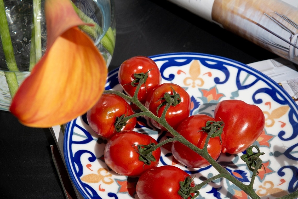 Una flor de cala naranja en primer plano desenfocado y un plato decorado con tomates cherry rojos en rama se presentan en una composición de naturaleza muerta, con elementos borrosos al fondo.