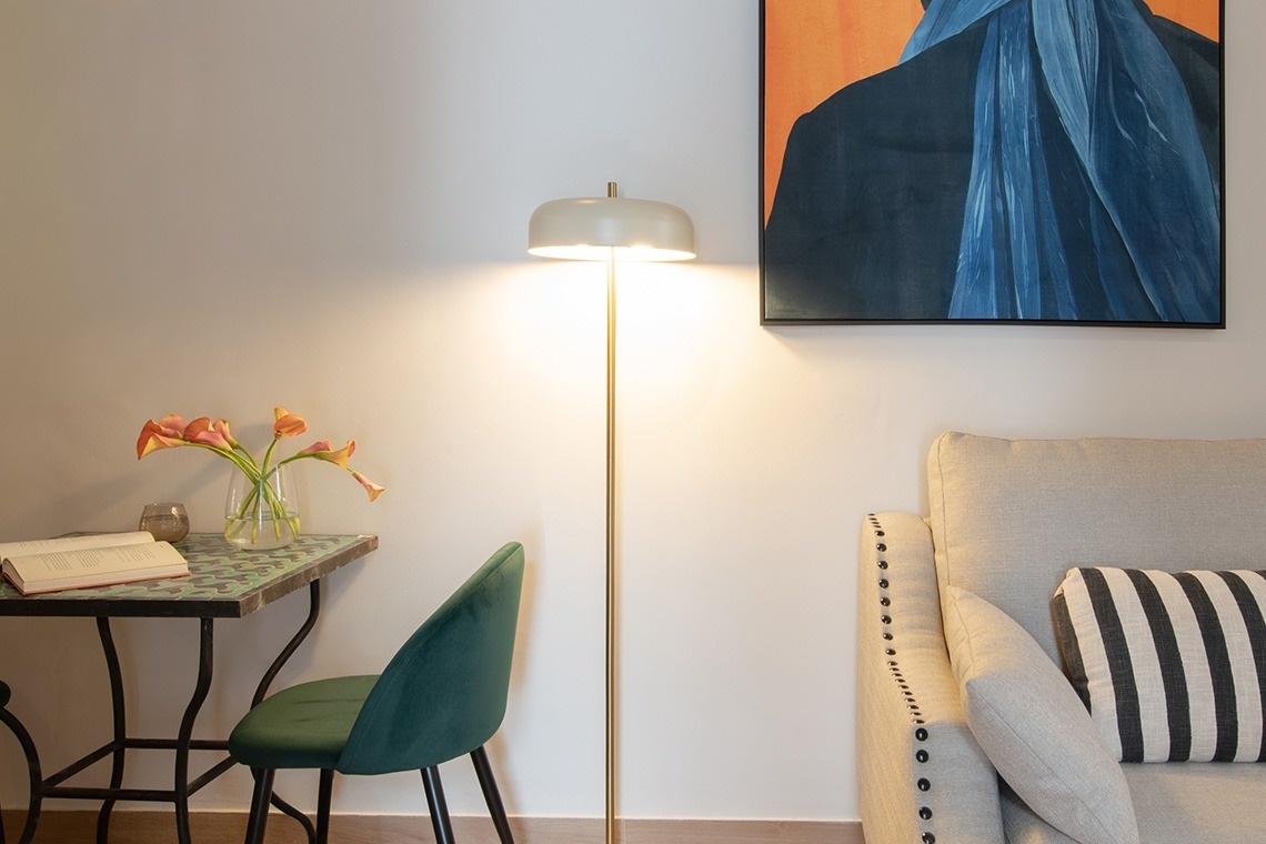 Una lámpara de pie ilumina un rincón acogedor de una sala con una mesa que tiene flores y un libro, una silla verde, parte de un sofá y un cuadro en la pared.