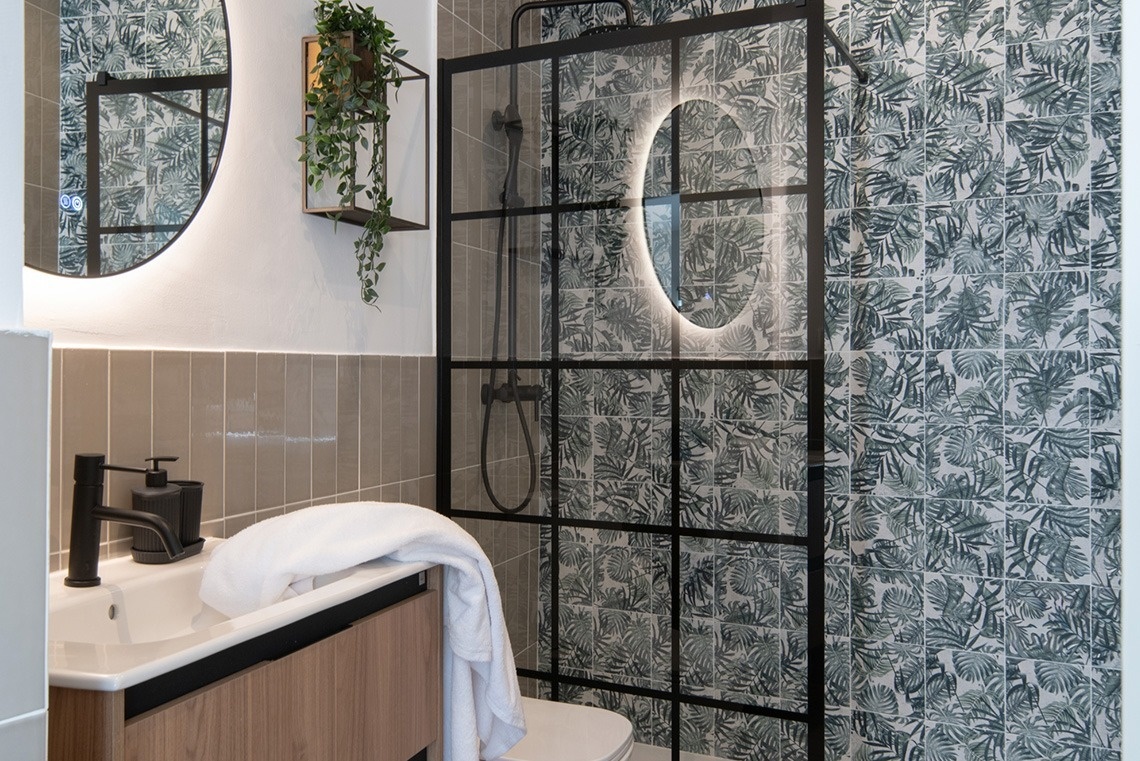 Un moderno baño presenta una ducha con mampara de rejilla negra y azulejos con estampado de hojas tropicales, junto a un tocador de madera con lavabo y un espejo redondo iluminado.