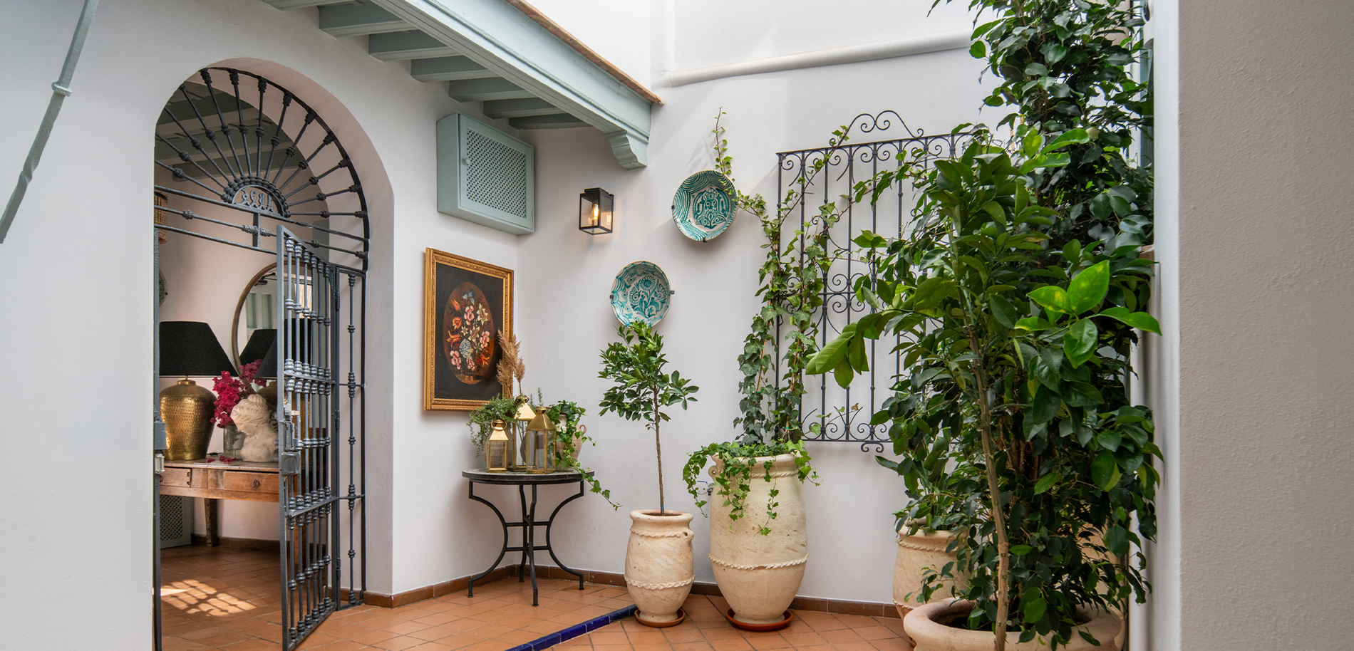 Un luminoso patio interior de estilo mediterráneo con paredes blancas, suelo de terracota, abundante vegetación en macetas y una elegante verja de hierro forjado que conduce a otra estancia.