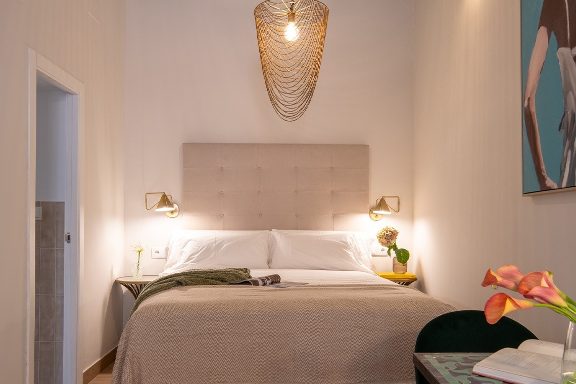 Una habitación luminosa y acogedora presenta una cama doble con un cabecero tapizado, lámparas de pared a cada lado y una llamativa lámpara colgante dorada, complementada con detalles florales y una pintura en la pared.