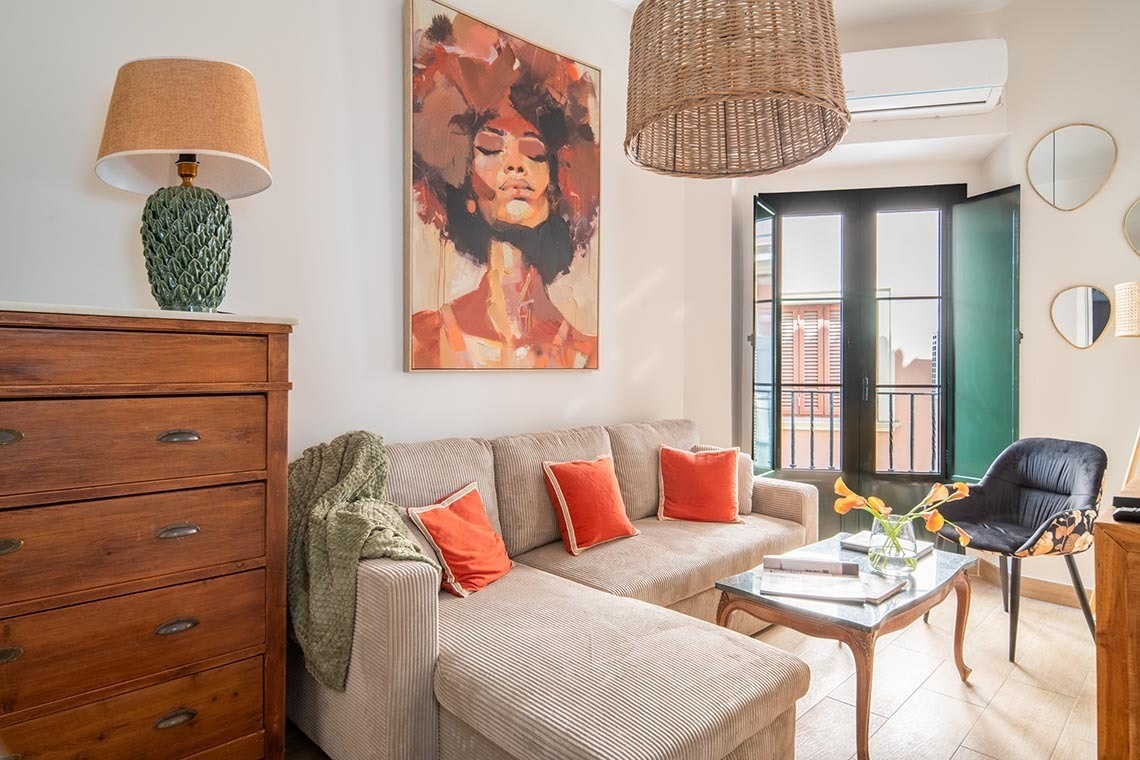 Un acogedor y luminoso salón presenta un sofá esquinero beige con cojines naranjas, una llamativa pintura de una mujer, una cómoda de madera y puertas de balcón con postigos verdes.