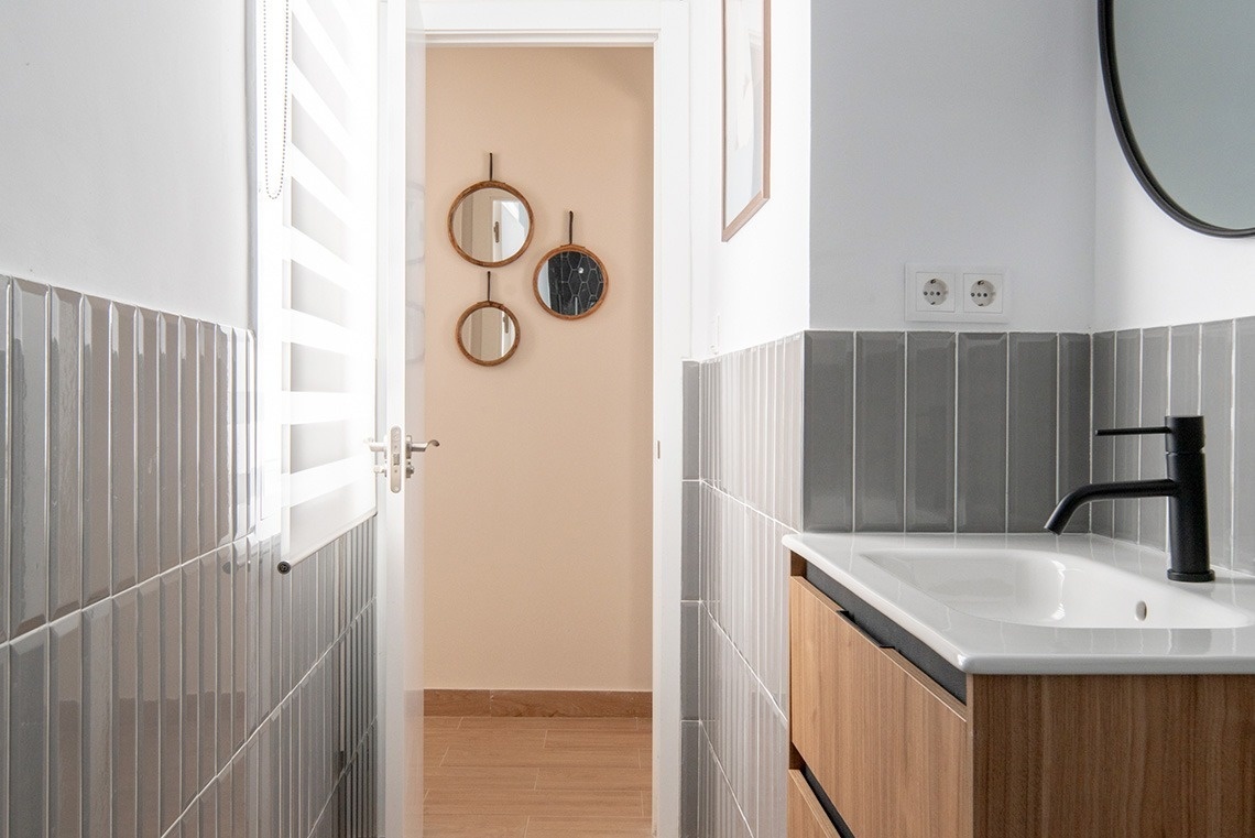 Moderno cuarto de baño con azulejos grises verticales, un lavabo con grifo negro sobre un mueble de madera, y una puerta que da a un pasillo decorado con espejos.