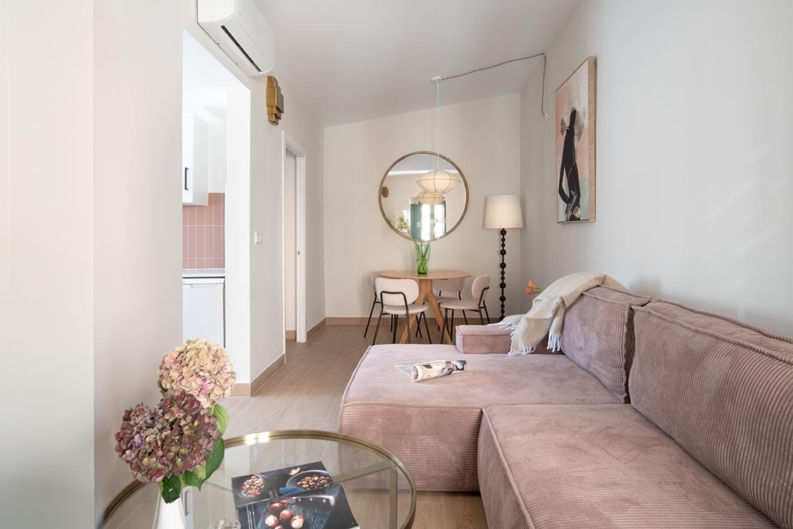 Una sala de estar moderna con un sofá modular rosa, un comedor con una mesa redonda y un espejo, y una cocina parcialmente visible al fondo.