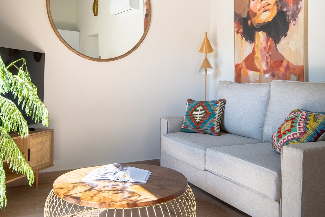 Una sala de estar luminosa presenta un sofá gris con cojines coloridos, una mesa de centro redonda de madera, un espejo circular y un cuadro artístico.