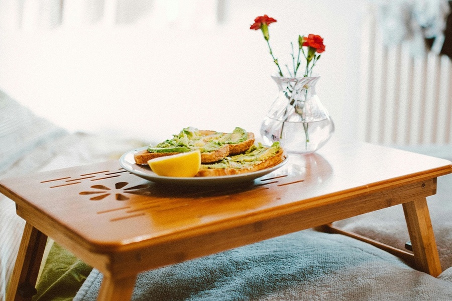 Un desayuno de tostadas de aguacate con un gajo de limón y un florero con flores rojas, presentado en una bandeja de madera sobre una cama.