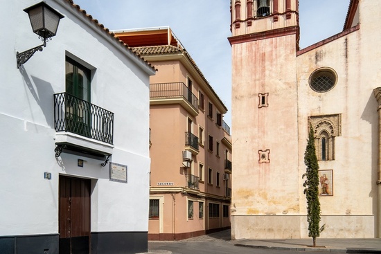 Una calle andaluza presenta una casa blanca, un edificio ocre y una iglesia antigua con torre, destacando sus distintas arquitecturas.