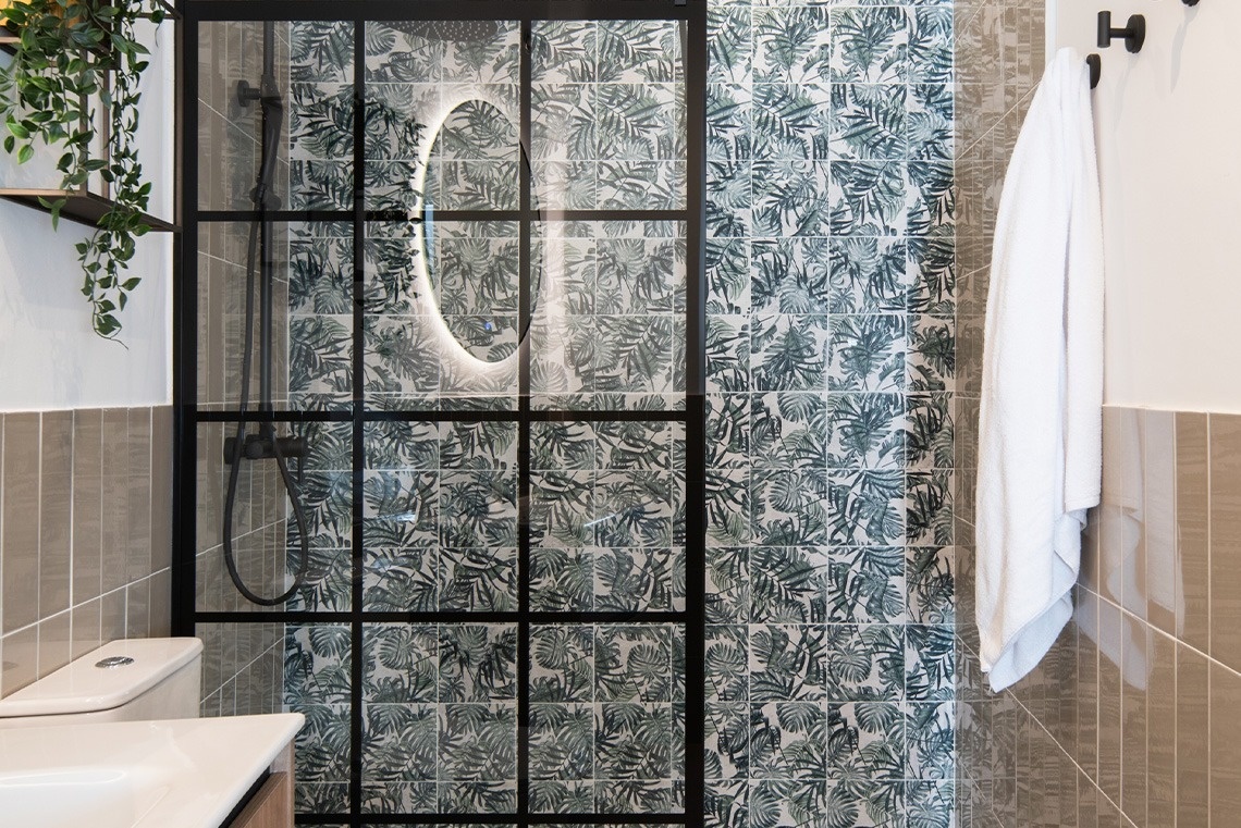 Este es un moderno cuarto de baño con una ducha de cristal con marco negro y azulejos de hojas tropicales, complementado con paredes de baldosas de tono neutro y un lavabo.