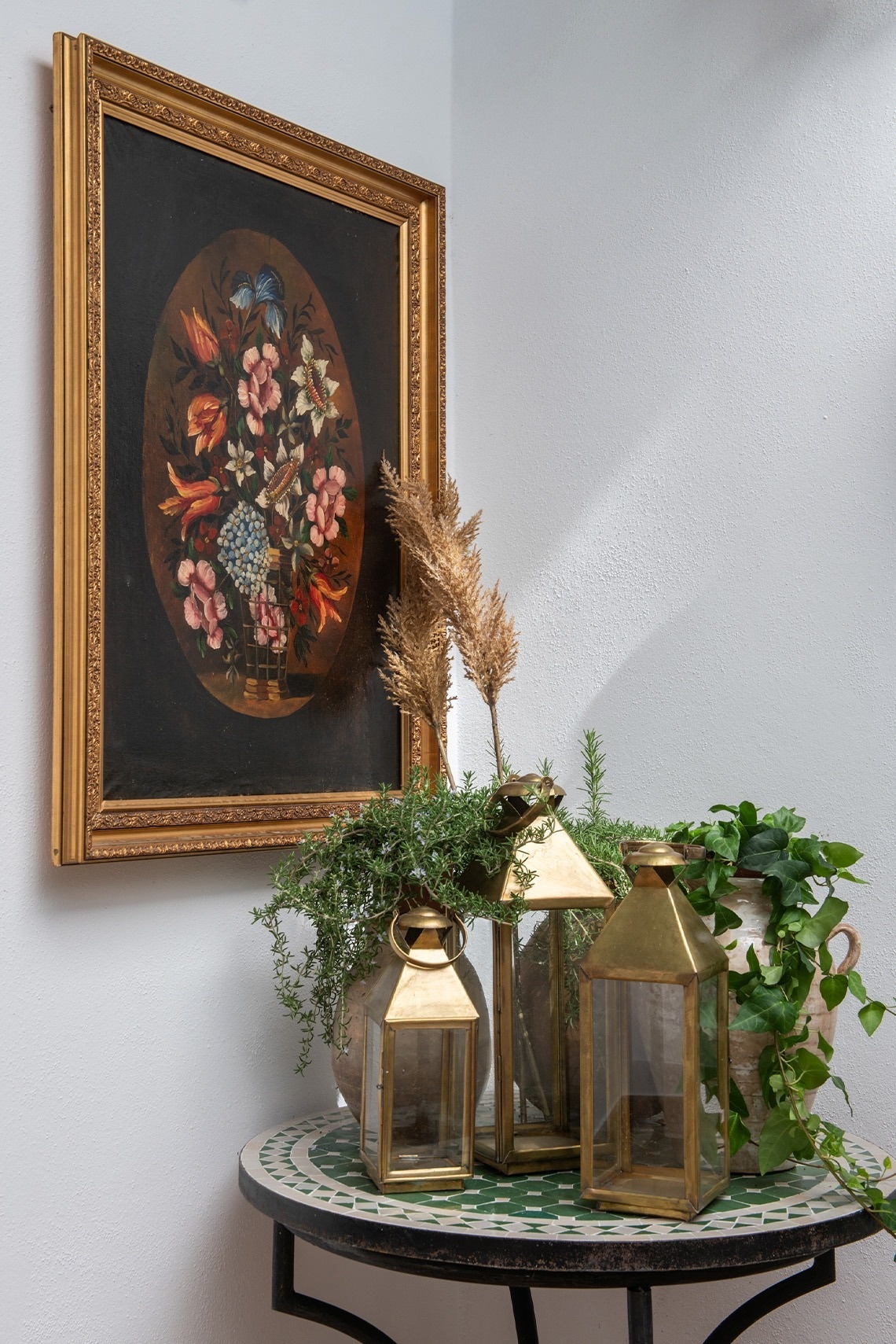 Una escena decorativa presenta un cuadro floral ovalado enmarcado en dorado, faroles de latón, plantas verdes y plumeros secos sobre una mesa redonda de mosaico.