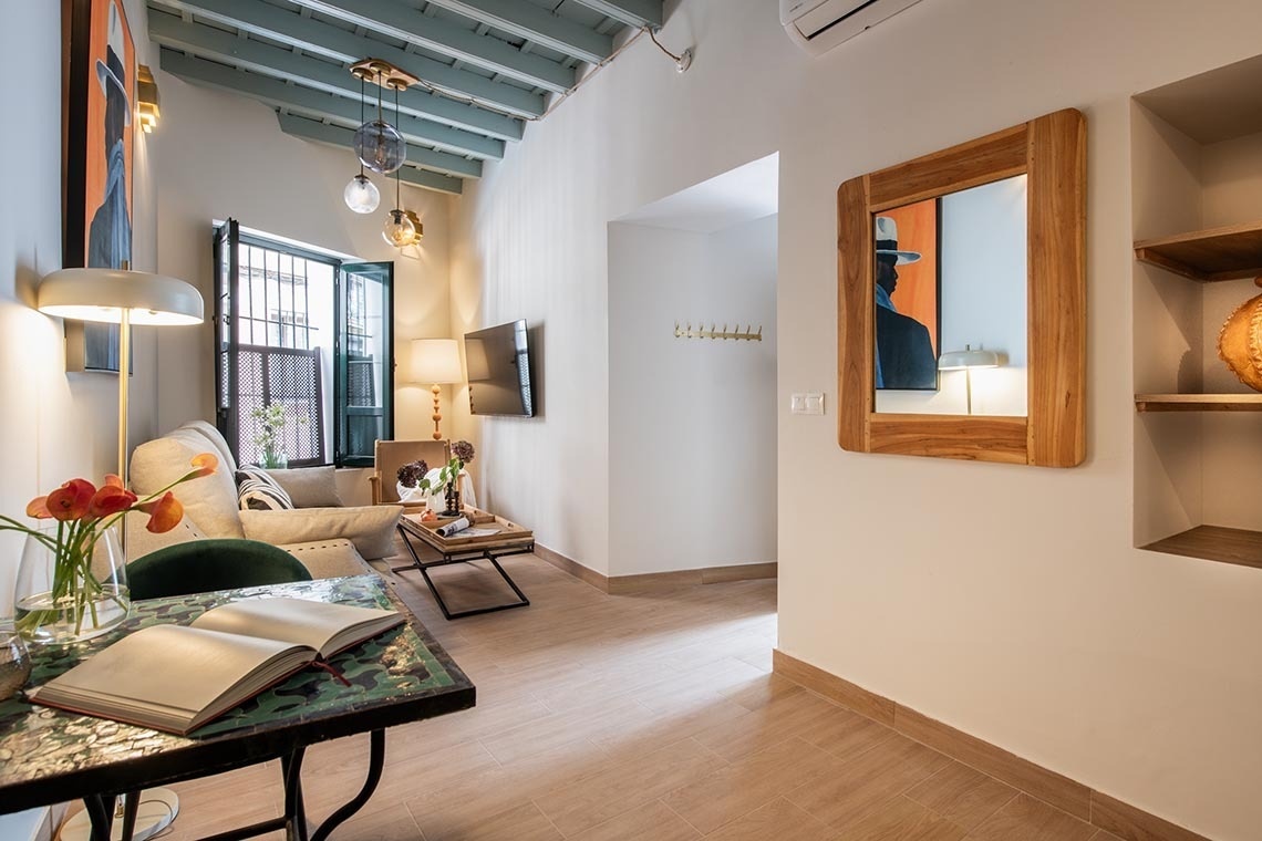 Un salón moderno y luminoso presenta un sofá, una mesa de centro con un libro y flores, un televisor, vigas vistas en el techo y un espejo de madera, creando un ambiente acogedor.