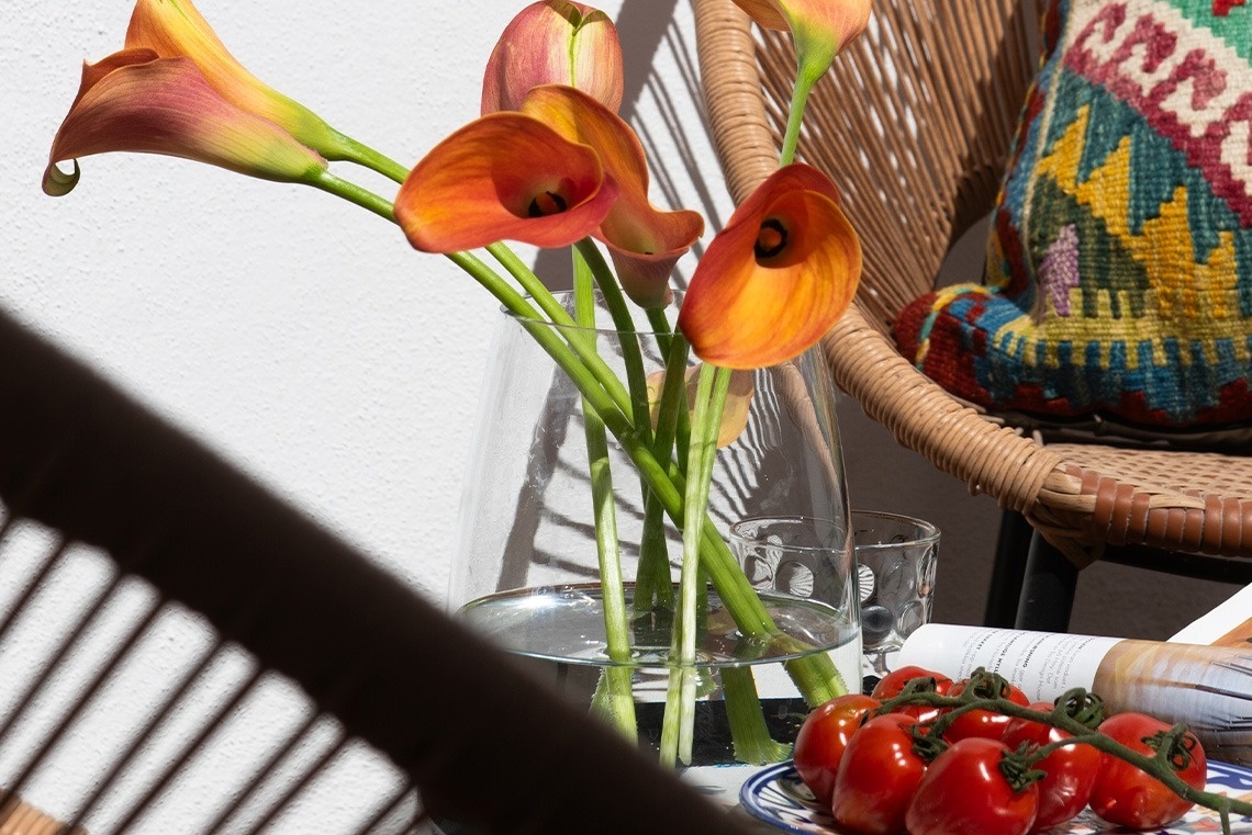 Un jarrón con calas naranjas y tomates cherry frescos se encuentran en una mesa junto a una silla de mimbre con un cojín colorido, con sombras rayadas proyectadas sobre una pared blanca.