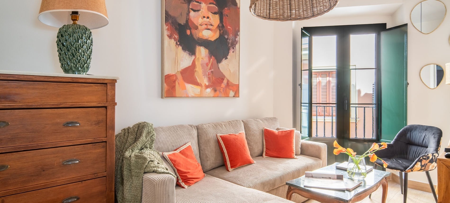 Un salón luminoso y acogedor presenta un sofá seccional beige con cojines naranjas, una llamativa pintura de una mujer en la pared, una cómoda de madera y un balcón abierto.