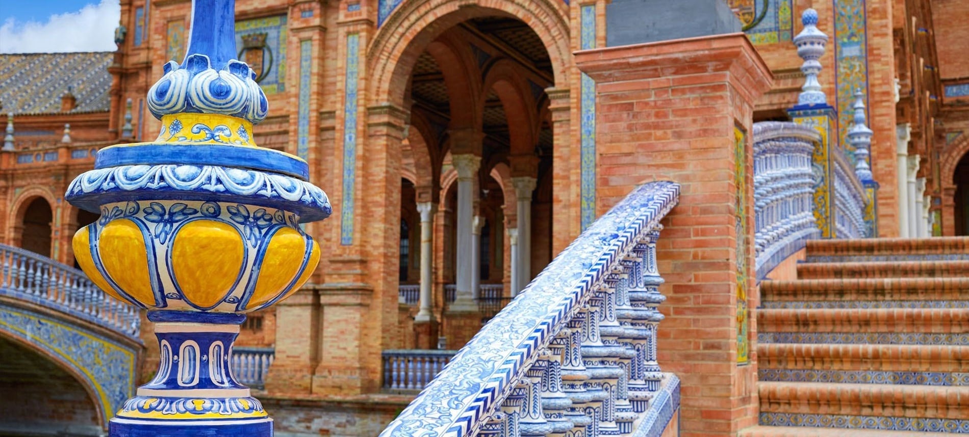 La imagen muestra intrincados detalles arquitectónicos con azulejos azules y amarillos en un pilar decorado, barandillas y escaleras, sobre un fondo de un edificio de ladrillo con arcos.