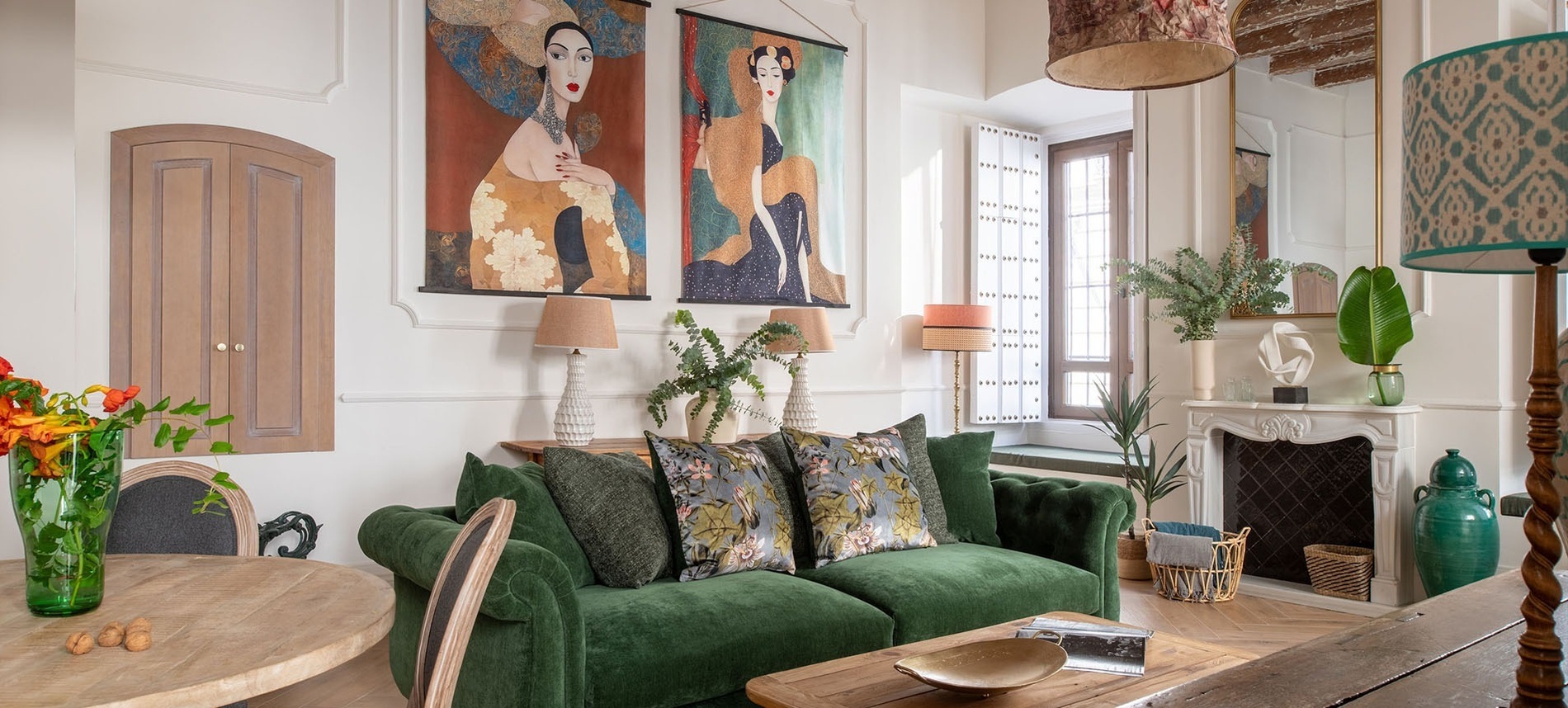 Un vibrante salón cuenta con un sofá de terciopelo verde esmeralda, grandes tapices artísticos de figuras femeninas y una cálida iluminación, creando un ambiente acogedor y ecléctico.