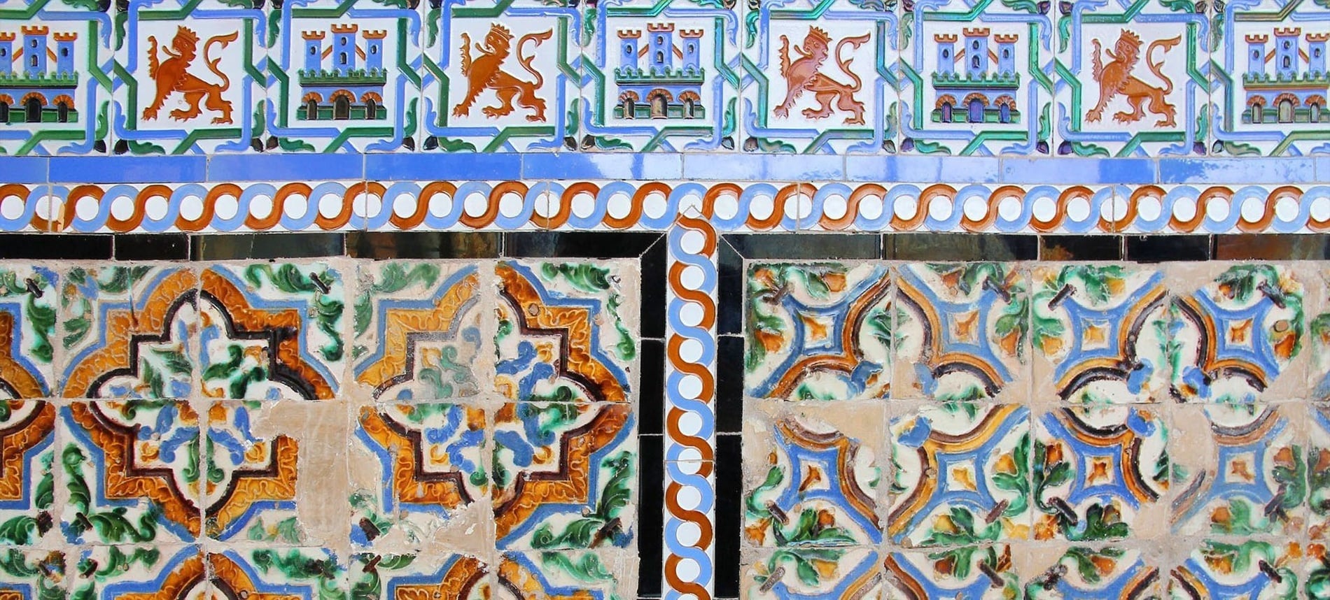 Una pared decorada con azulejos cerámicos de intrincados diseños en tonos azules, naranjas, verdes y blancos, presentando motivos heráldicos de leones y castillos, junto con patrones geométricos y florales.