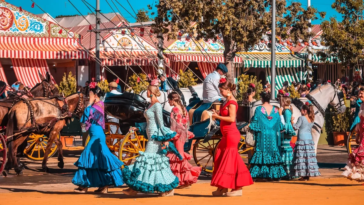 Women in vibrant flamenco dresses and horse-drawn carriages animate a bustling Spanish fairground, complete with colorful striped stalls under a clear blue sky.