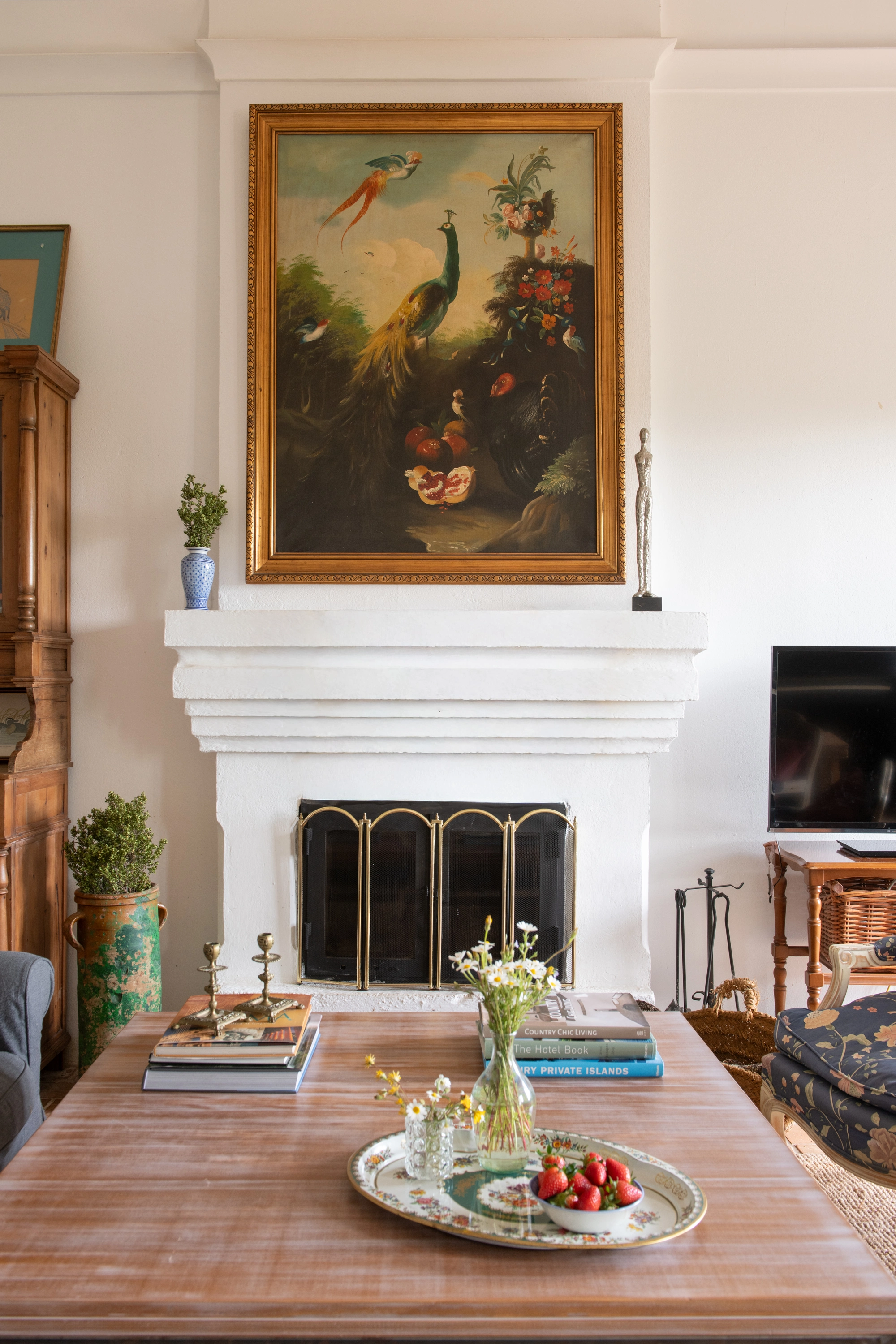 Elegant living room with painting above fireplace, a coffee table with strawberries and books, and a view of a chair Epoca Suites: Corporativa.