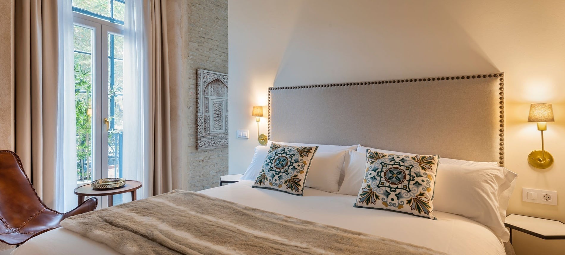 Un dormitorio acogedor y elegantemente decorado presenta una cama hecha con cojines bordados, un cabecero tapizado, apliques de pared y una luminosa ventana.