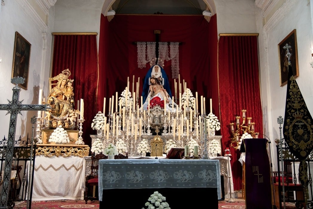 Un altar de iglesia ricamente adornado presenta una Piedad central rodeada de un mar de velas y flores blancas, con cortinas rojas de fondo.