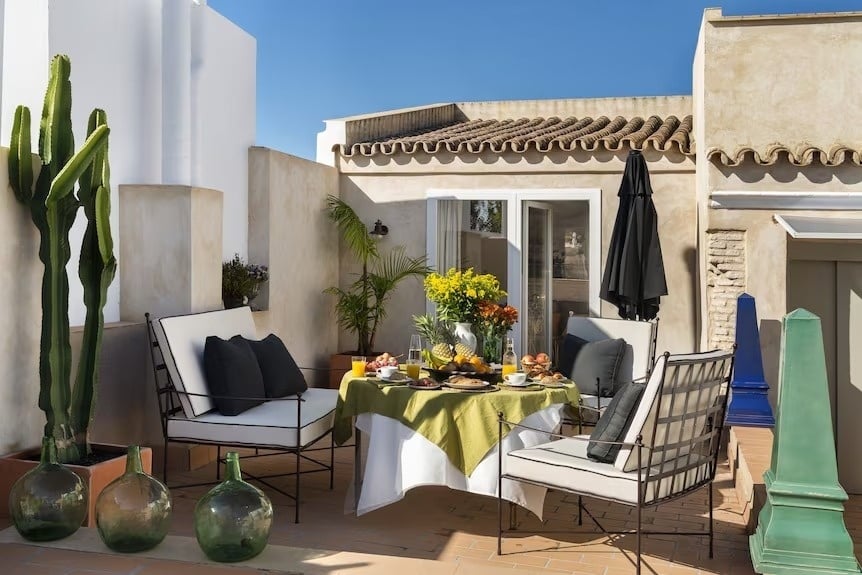 A beautifully set outdoor patio, featuring a table laden with breakfast and flowers, is surrounded by comfortable seating, various plants, and traditional Mediterranean-style architecture under a clear blue sky.