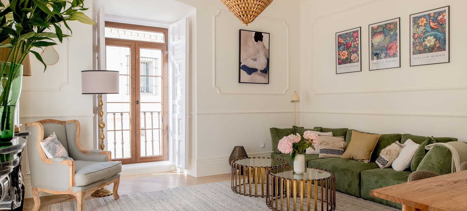 Un luminoso salón de estilo clásico y moderno combina un sofá verde con cojines, un sillón vintage, y cuadros decorativos en las paredes blancas con molduras, junto a un gran ventanal de madera.