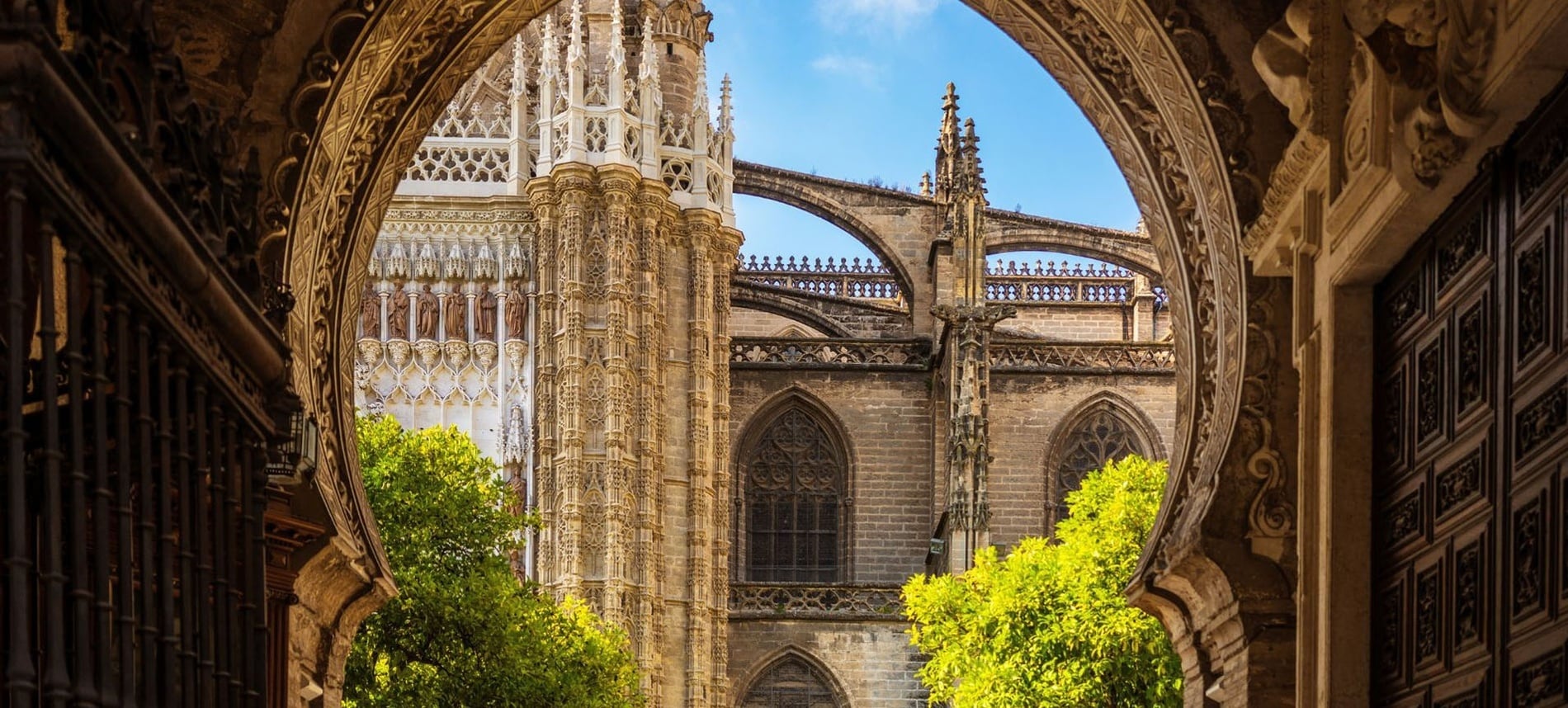 Un arco ricamente ornamentado de influencia morisca encuadra una vista de la elaborada arquitectura gótica de una catedral, destacando sus contrafuertes y pináculos bajo un cielo azul y entre árboles verdes.