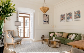A bright and elegantly furnished living room features a green sofa, metallic coffee tables, and natural light streaming in from a large window with a balcony.