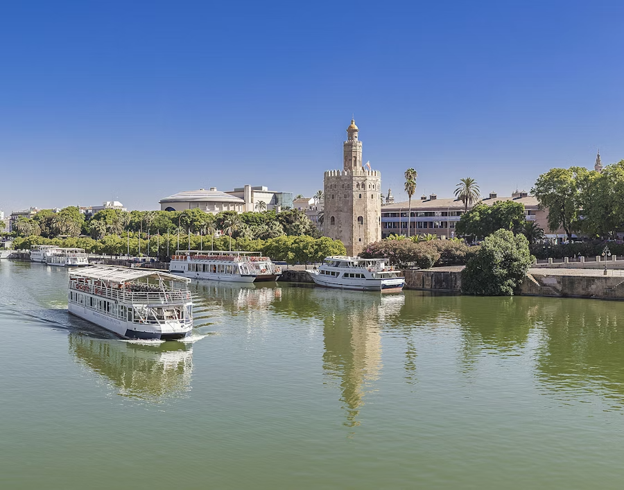 Several tour boats navigate a calm, green river, reflecting a historic tower (Torre del Oro) and a tree-lined cityscape under a clear blue sky.