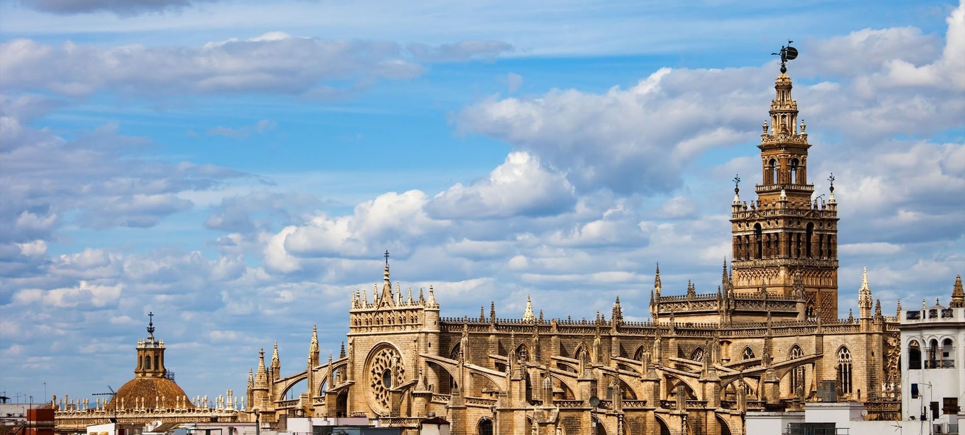 La majestuosa Catedral de Sevilla y su icónica torre Giralda se alzan prominentes bajo un cielo azul salpicado de nubes blancas.