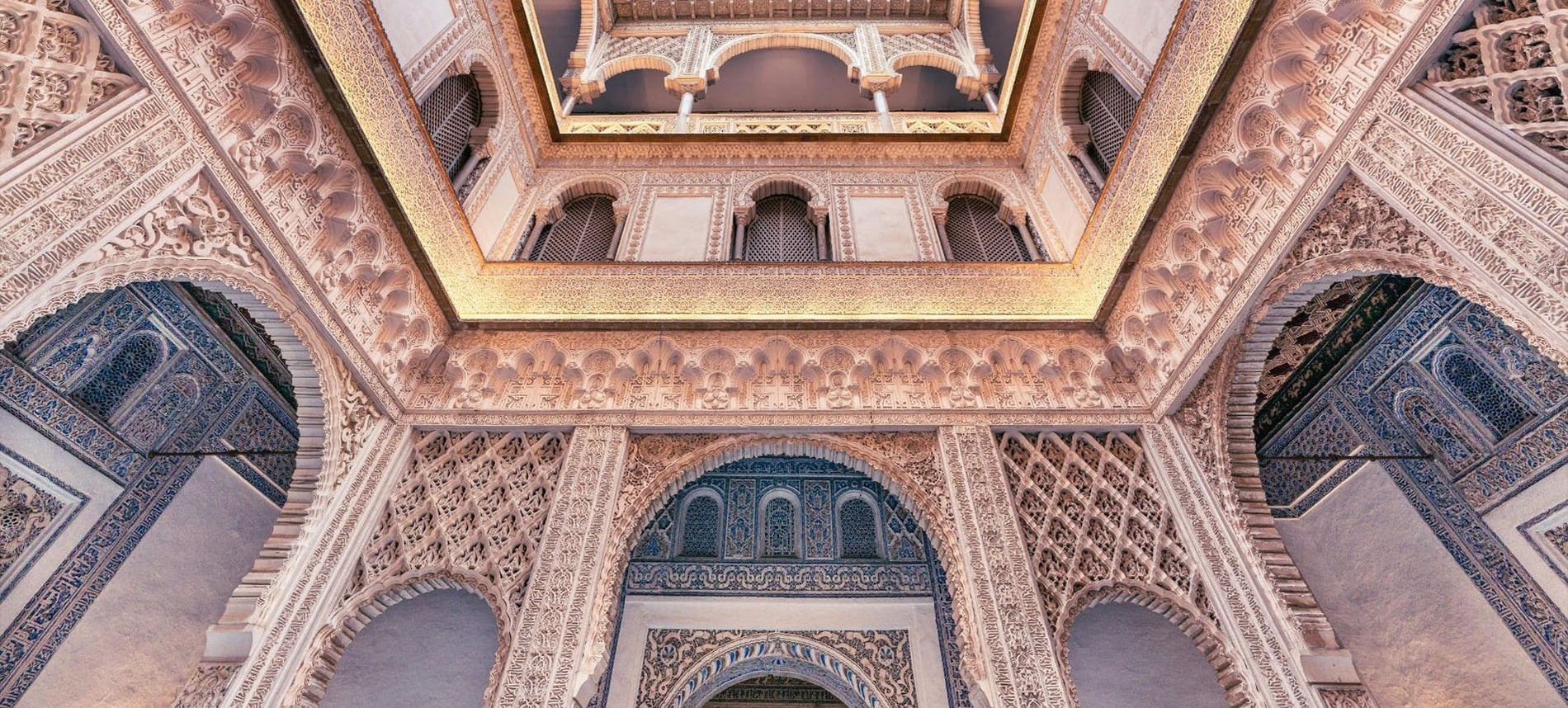 Detalle ascendente de un patio interior de estilo mudéjar, ricamente decorado con intrincados tallados, arcos y azulejos azules.