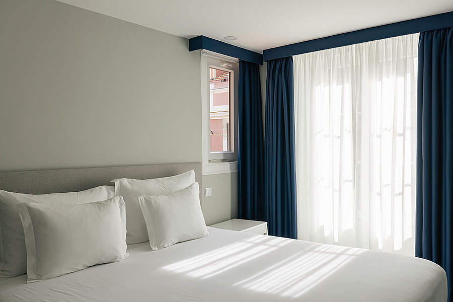Cama confortável em quarto de hotel com cortinas azuis e brancas, e janela com vista Esqina: Corpo