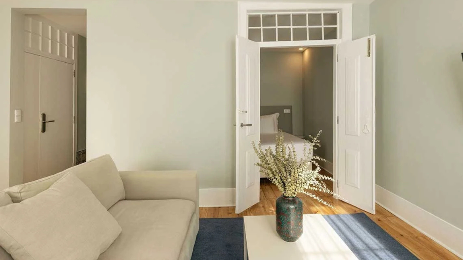Cozy hotel room with a sofa, open doors to a bedroom, and a vase of flowers. Esqina: Corpo