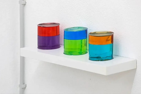 tres vasos de vidrio de colores están sentados en una estantería blanca .