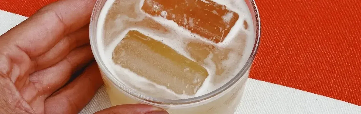 Um copo de bebida com gelo, segurado por uma mão, sobre uma toalha de mesaEsqina: Cosmopolitan listrada em vermelho e branco.