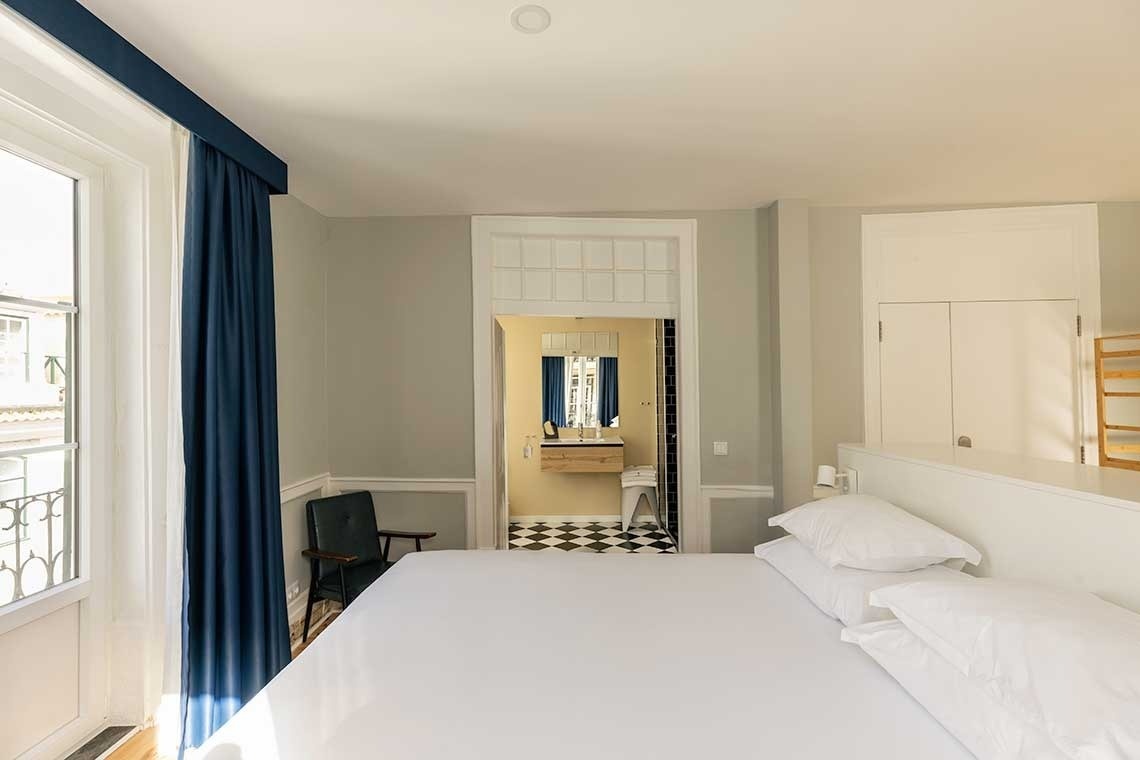 Una vista de un dormitorio con cama, ventana y cortinas azules, abriéndose a un baño con suelo de baldosas a cuadros.