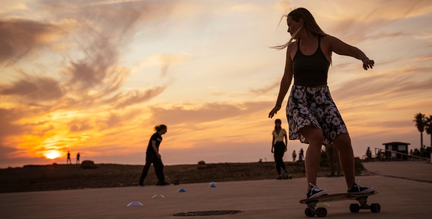 a woman in a floral skirt is riding a skateboard at sunset