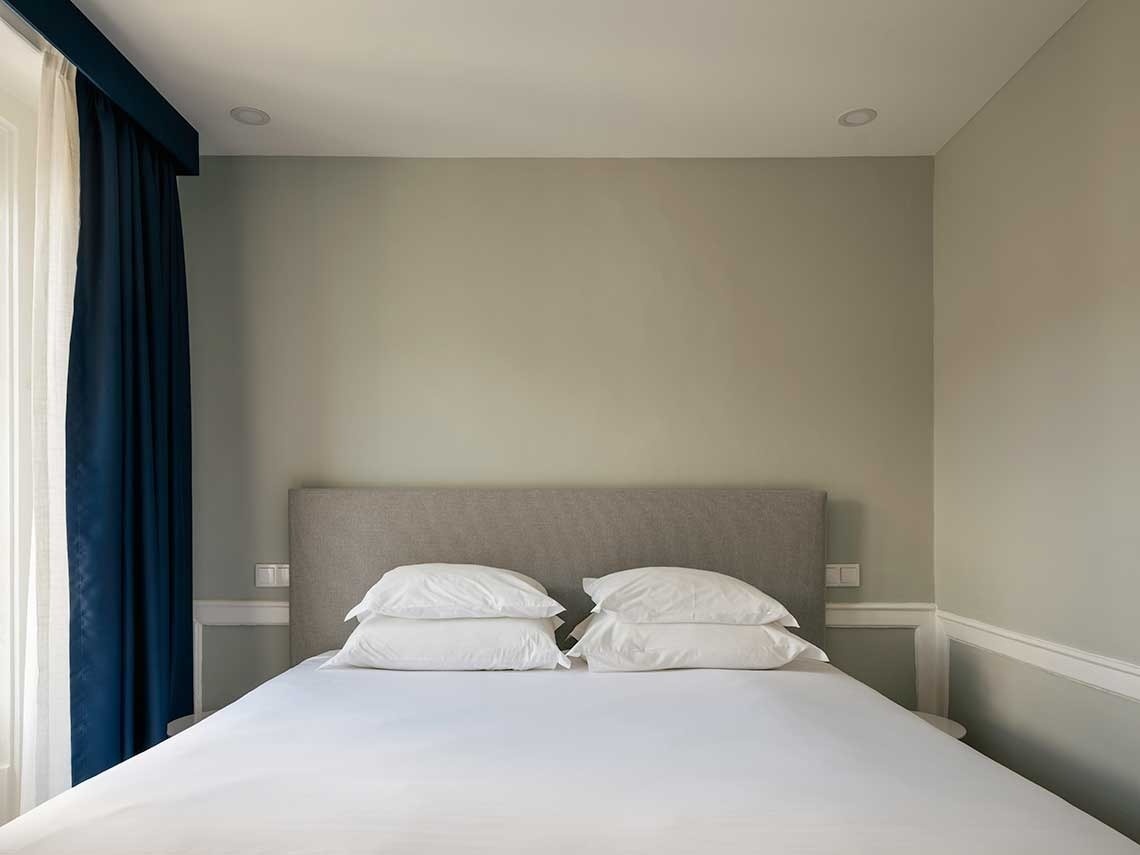 Una moderna habitación de hotel presenta una cama hecha con cabecero gris y sábanas blancas, paredes de color verde claro y cortinas azul oscuro que cuelgan junto a una ventana.