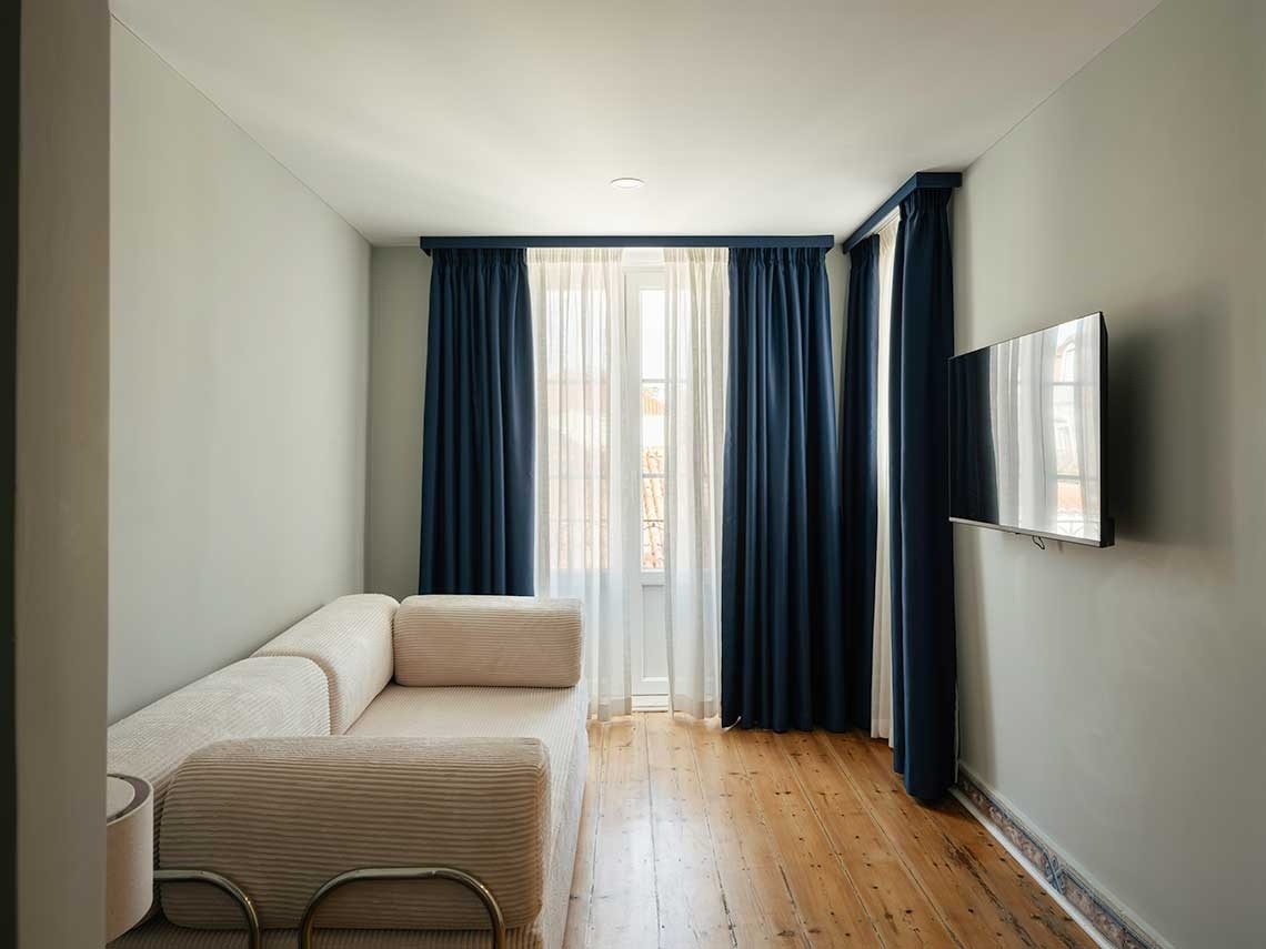 Salón con paredes verde pálido, suelo de madera, un sofá beige acanalado, cortinas azul marino en la ventana y un televisor en la pared.