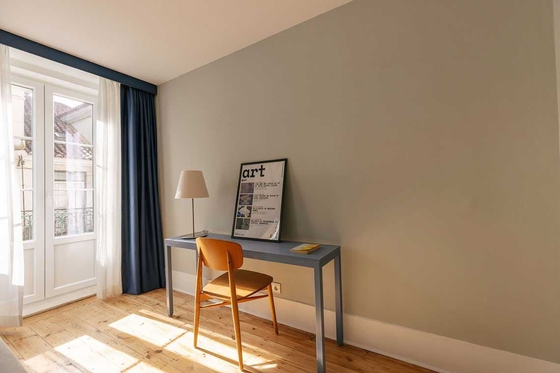 A modern room features a simple desk with a lamp and an "art" poster, a light wooden chair, and a large window with white and blue curtains, all set on a sun-dappled wooden floor.