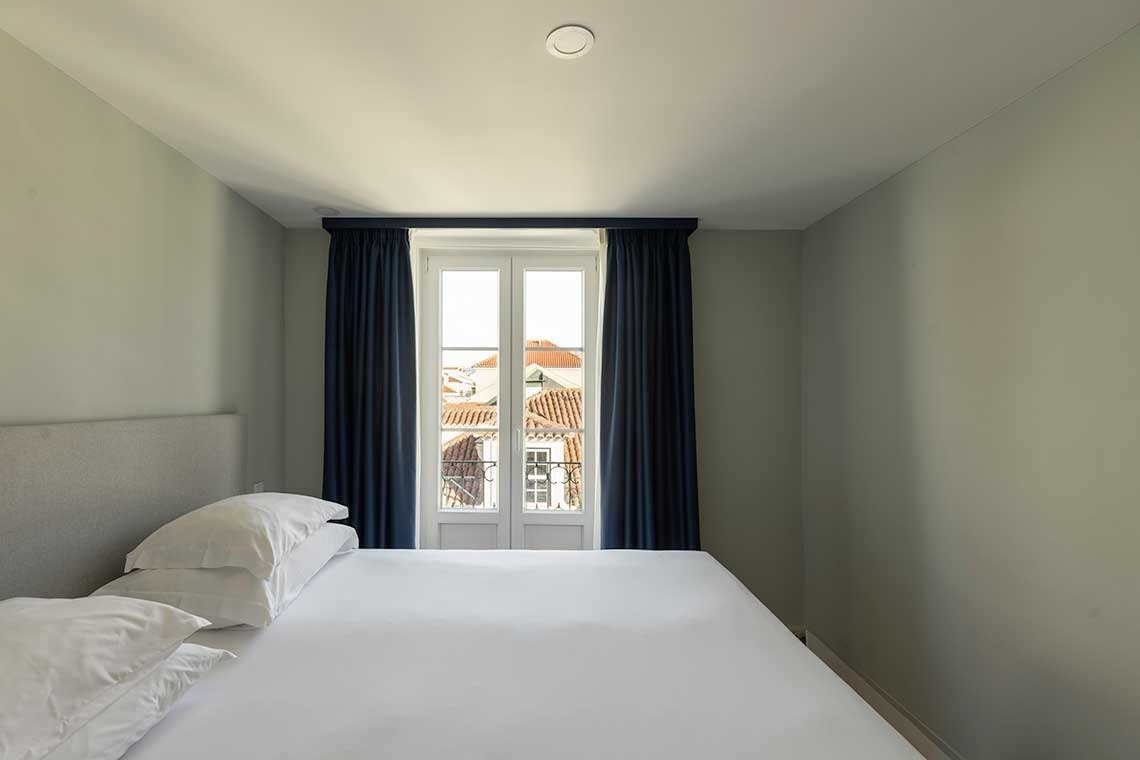 Un dormitorio luminoso con una cama blanca, paredes gris claro y una ventana central con cortinas azules que ofrece vistas a tejados de terracota.