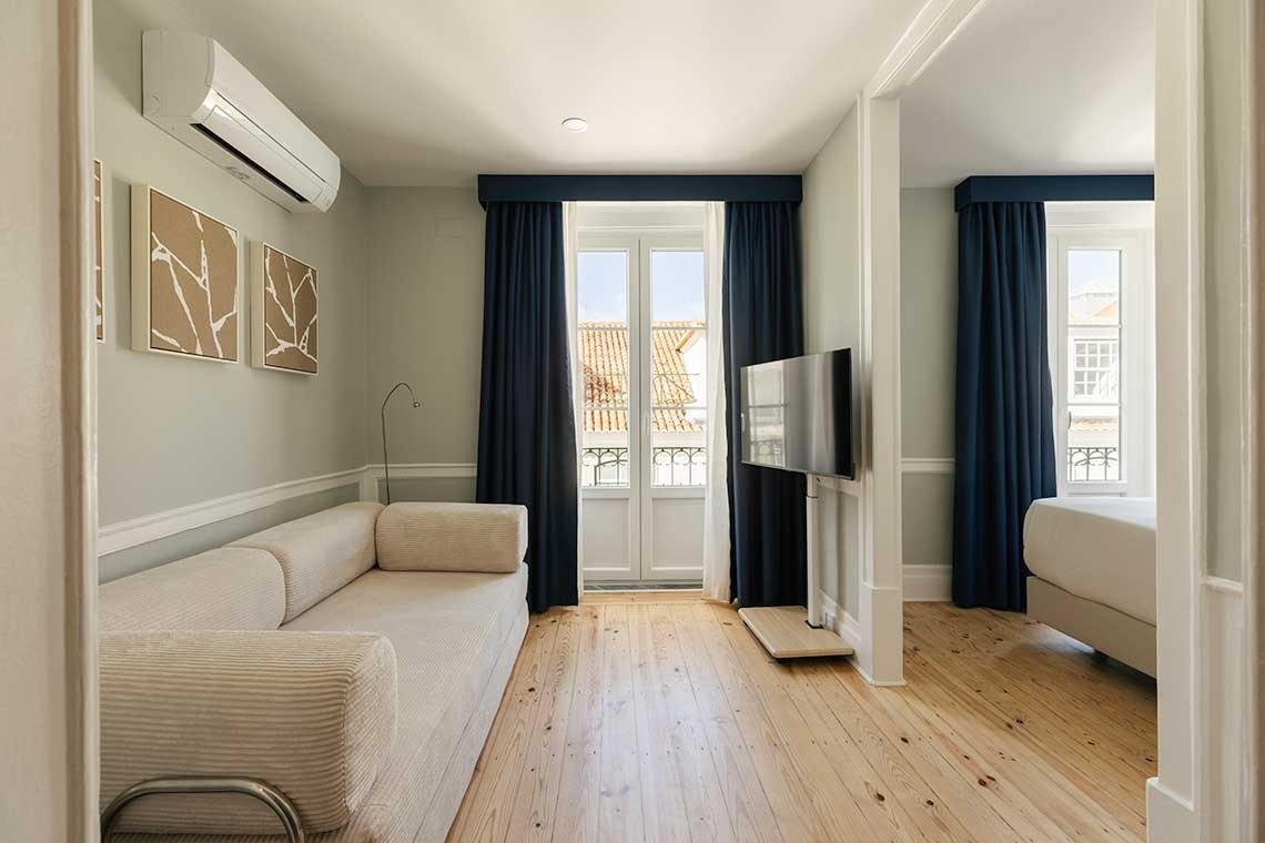 Una luminosa sala de estar con un sofá beige, aire acondicionado, televisión y grandes ventanales con cortinas azules se abre a un dormitorio adyacente, todo sobre un suelo de madera clara.