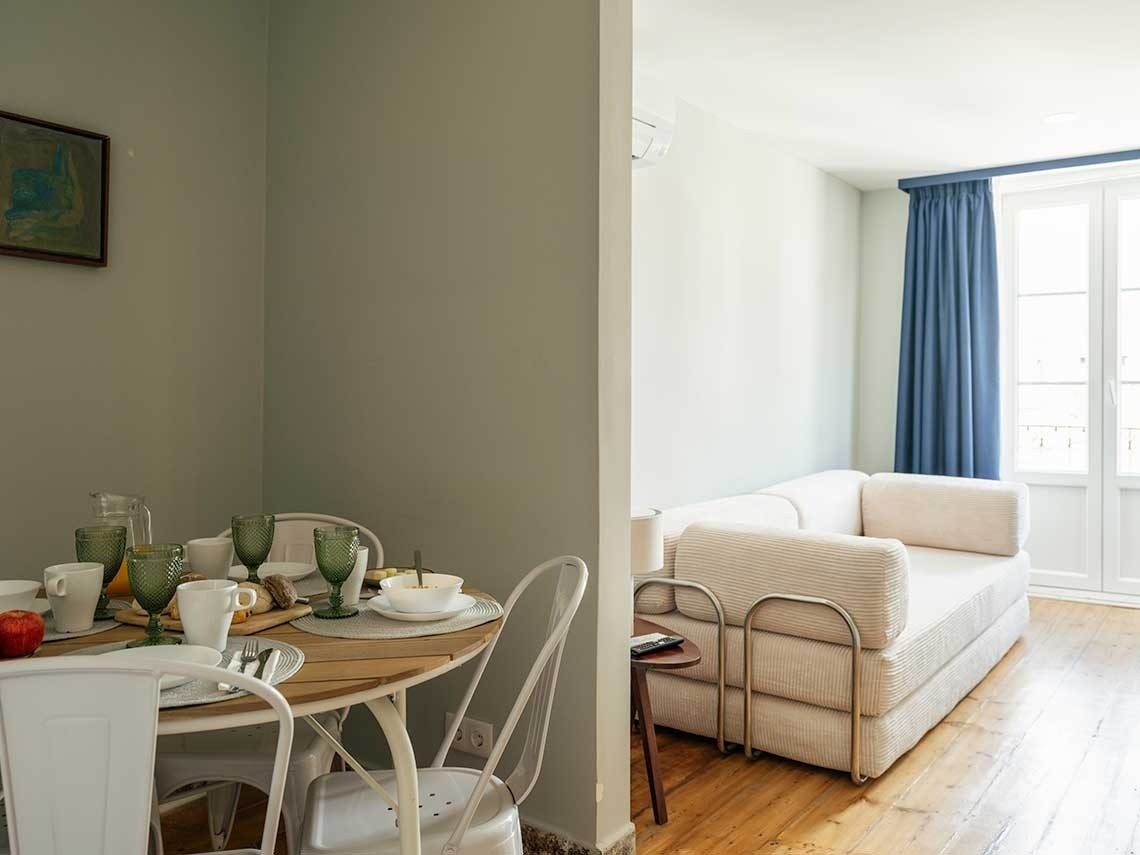 La imagen muestra un espacio interior dividido, con un comedor con mesa puesta y sillas blancas en primer plano, y una sala de estar con un sofá cama y cortinas azules visible al fondo.