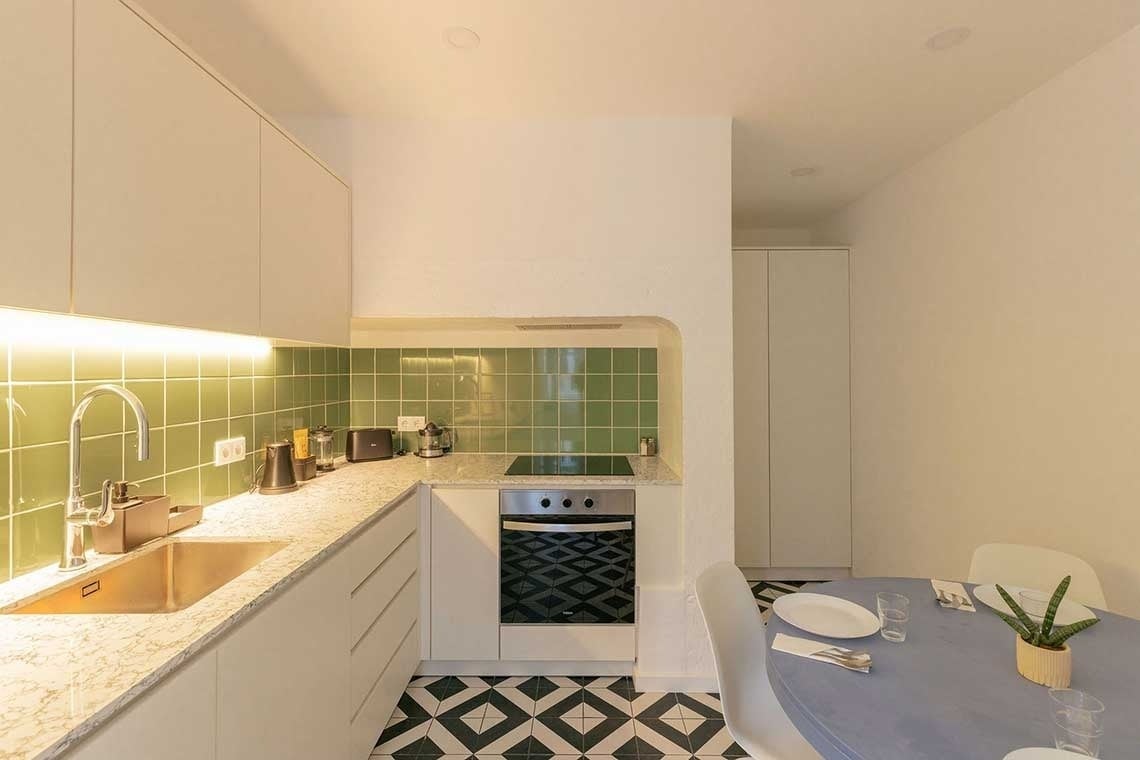 A stylish kitchen, boasting white cabinets, a green tiled backsplash, and monochromatic patterned flooring, seamlessly transitions into a cozy dining area with a blue table.