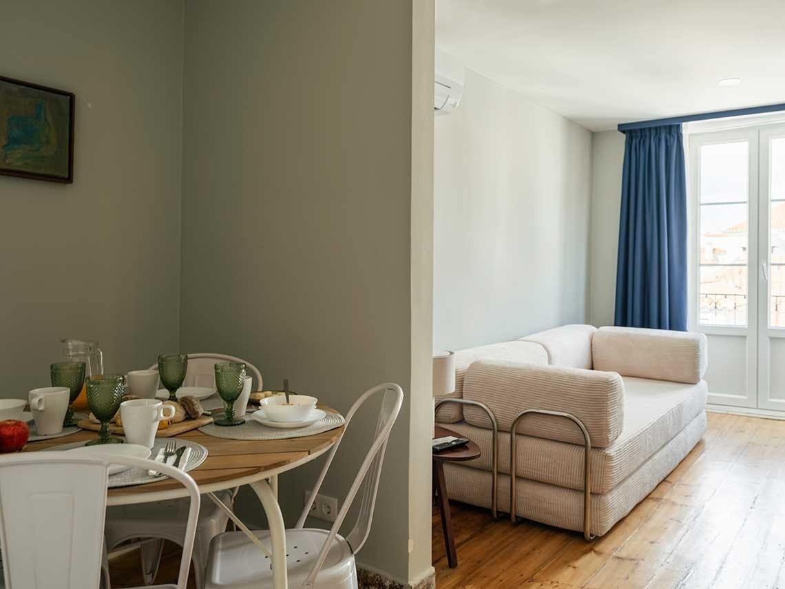 Una imagen interior que muestra un área de comedor con la mesa puesta para el desayuno, adyacente a un área de estar con un sofá y una ventana con cortinas azules.