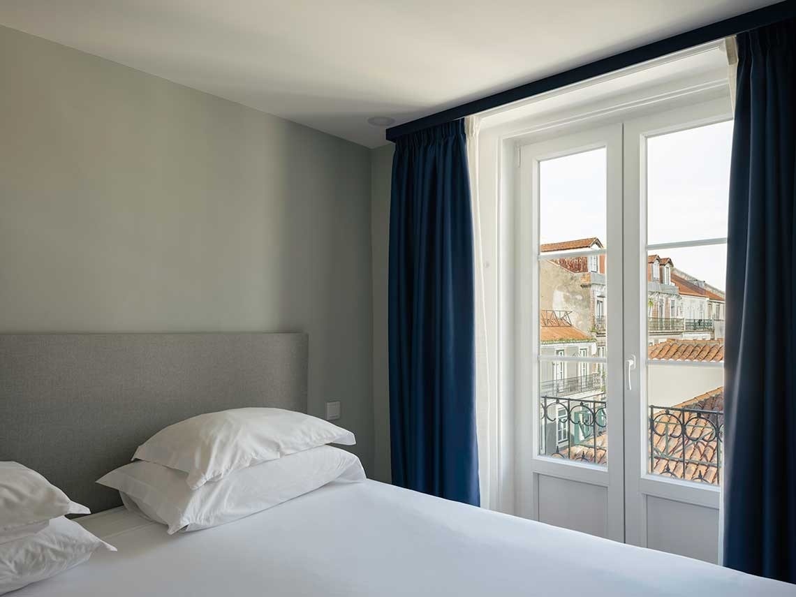 Un dormitorio moderno y luminoso con una cama blanca, almohadas apiladas y un gran ventanal con cortinas azul marino que da a edificios antiguos con tejados de terracota.