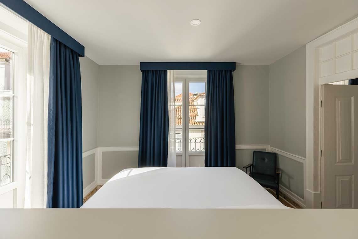 Una habitación luminosa presenta una cama con sábanas blancas, paredes gris claro con molduras blancas, cortinas azules en dos ventanas y una puerta blanca a la derecha, con un atisbo de tejados exteriores.