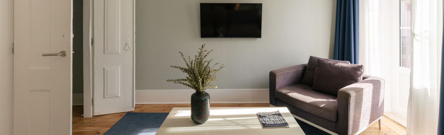 Un dormitorio en escala de grises presenta una cama hecha, un televisor, una silla y una ventana con vista a un edificio.