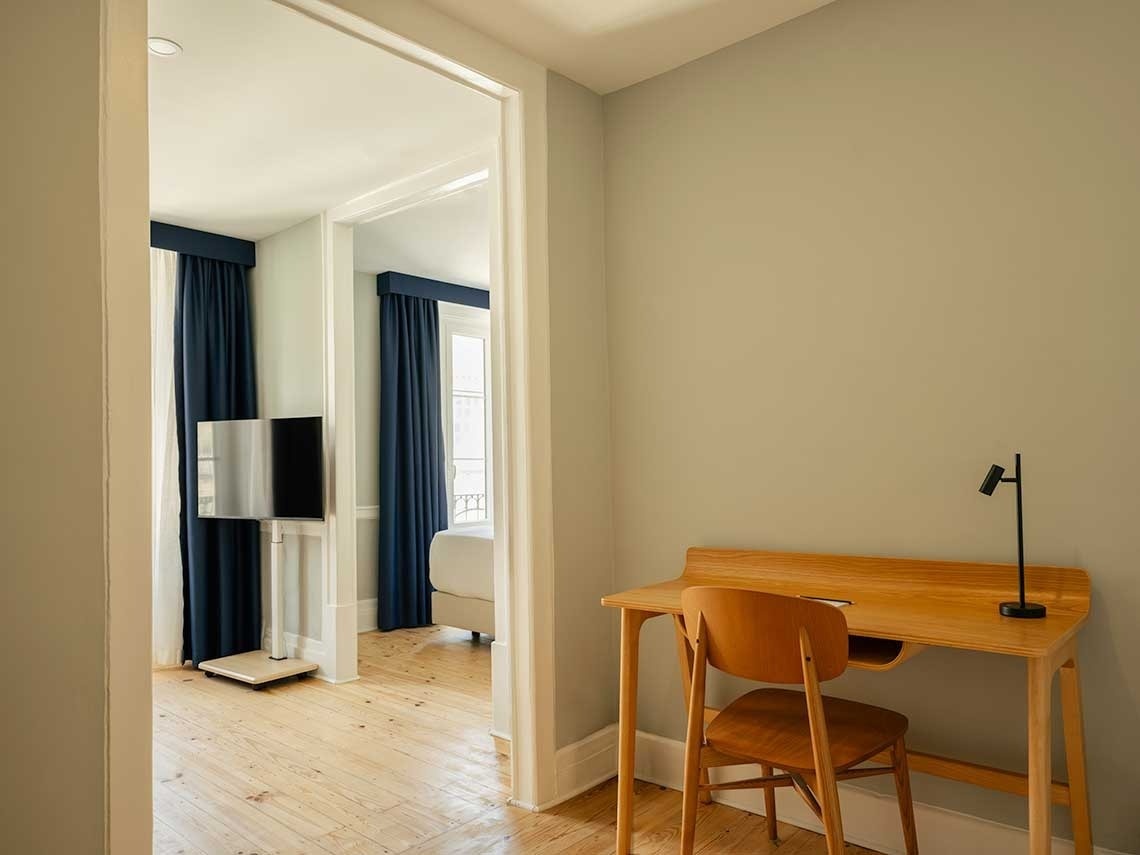 La imagen muestra una habitación con un escritorio y una silla de madera en primer plano, y a través de una puerta abierta, se vislumbra otra habitación con un televisor de pantalla plana y cortinas azules.