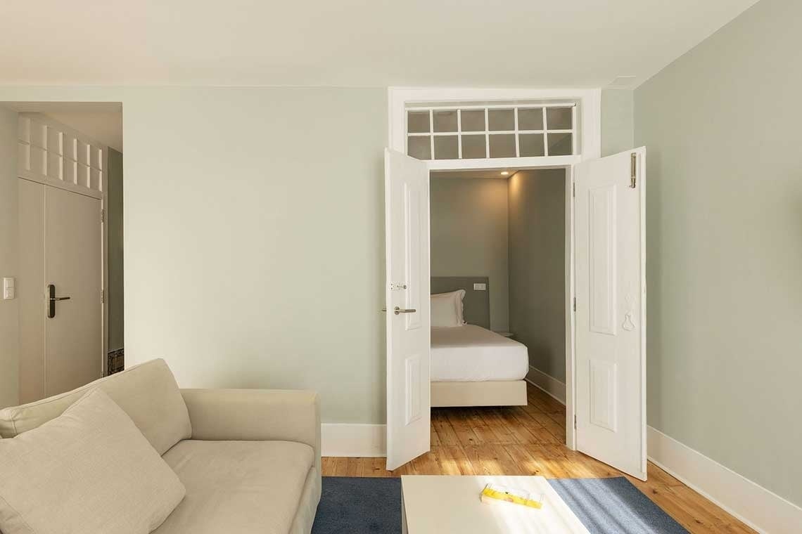 A bright, minimalist living room with a cream sofa and a coffee table features an open double doorway leading into a cozy bedroom with a white bed, under a transom window.